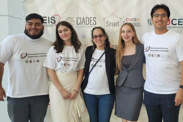 Four students pose with Professor Karla Badillo-Urquiola for a photo in front of a banner for the International School on Responsible Computing.