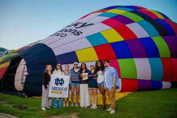 Seven students stand on grass in front of a partially inflated, colorful hot air balloon. They hold a banner reading "ND Mexico Club".  The balloon displays a checkered pattern of red, yellow, green, blue, purple, and white sections.
