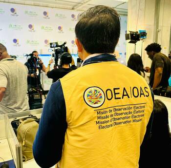A person wearing a yellow OEA/OAS Electoral Observation Mission vest stands facing several videographers at an event. Behind them, a white wall displays repeated TRE-SP logos.