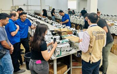 Personnel from an Electoral Observation Mission prepare white electronic voting machines in a large room. Multiple people, including a man in a beige OEA/OAS vest writing on a clipboard, work at tables lined with machines and cables.