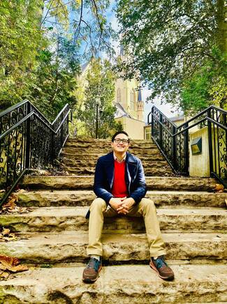 A smiling man with glasses, wearing a navy coat and red sweater, sits on rugged stone steps with black railings. The Basilica of the Sacred Heart rises behind autumn trees in the background.