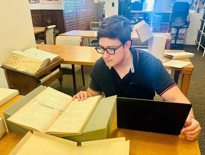 Dark-haired man in glasses and a dark polo shirt intently examines an open ledger book with handwritten script, with a laptop nearby in a university archives research room.