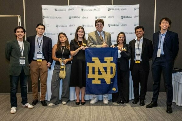 Eight smiling individuals, five men and three women, in business casual attire, proudly hold a large blue and gold Notre Dame flag before a repeating University of Notre Dame Mexico logo backdrop. One man gives a thumbs up.