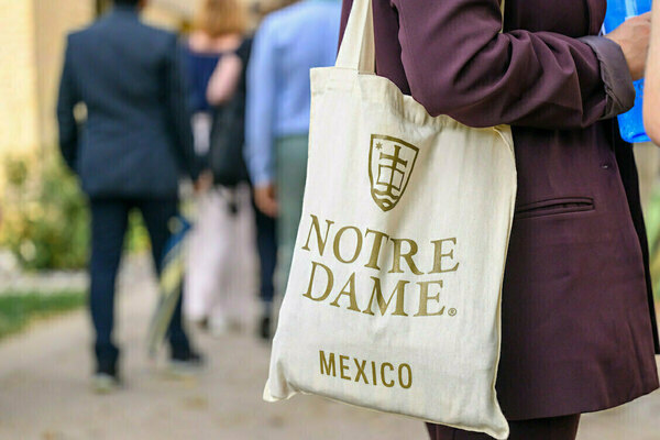A person in a burgundy blazer carries a natural canvas tote bag featuring the gold Notre Dame logo and "NOTRE DAME MEXICO" text, walking past blurred people outdoors.