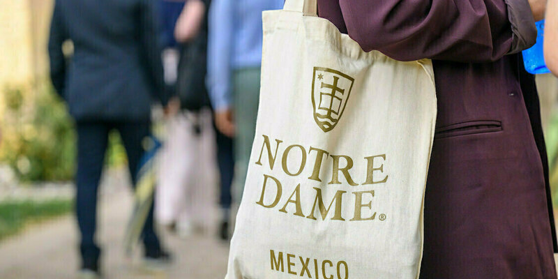A person in a burgundy blazer carries a natural canvas tote bag featuring the gold Notre Dame logo and "NOTRE DAME MEXICO" text, walking past blurred people outdoors.