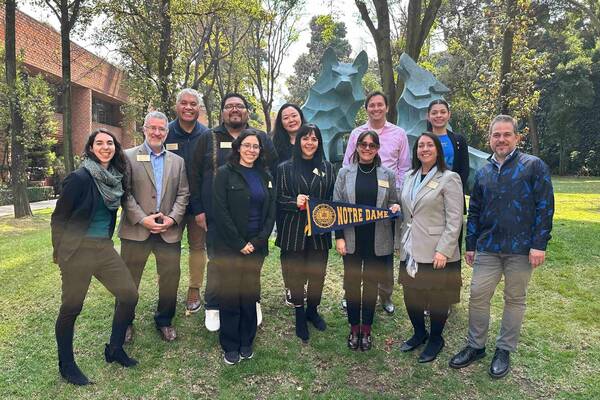 Eleven smiling people, some with nametags, pose outdoors with a blue Notre Dame pennant and an abstract teal wolf sculpture.