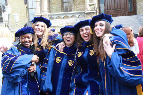 Five joyful graduates in blue velvet and gold academic regalia, embracing and smiling in front of a Notre Dame campus building.