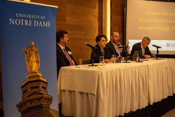 Three men and one woman speak at a table with microphones. A blue Notre Dame banner features the Golden Dome.