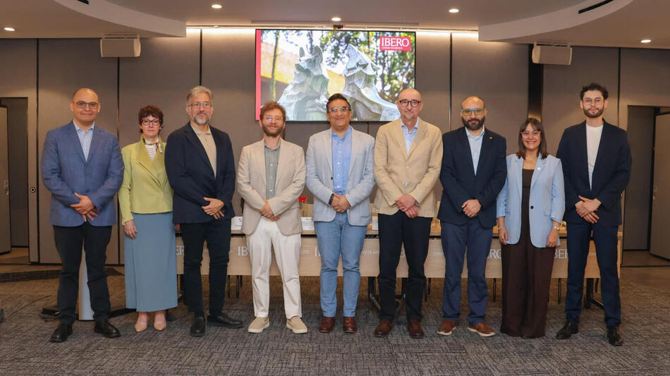 Nine people in business casual attire smile, standing before a screen displaying 'IBERO CIUDAD DE MÉXICO'.