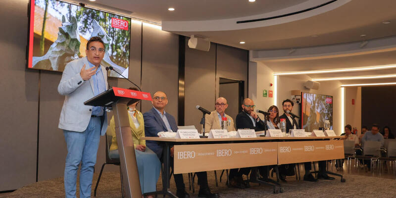 Speaker in light blue blazer gestures at podium. Six panelists sit at a table with IBERO logo, screen displays IBERO Ciudad de México.
