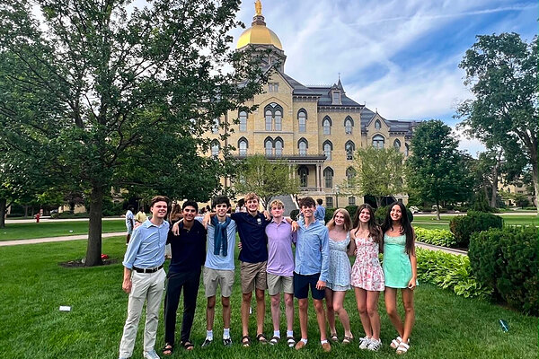 Nine smiling young adults pose on green grass with arms around each other, Main Building and Golden Dome in background.