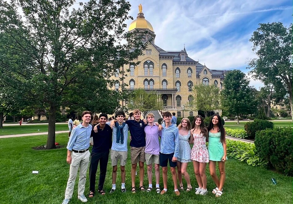 Nine smiling young adults pose on green grass with arms around each other, Main Building and Golden Dome in background.