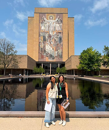 Two smiling young women stand before the Hesburgh Library's "Word of Life" mural and reflecting pool.