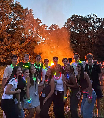 A group of smiling students wearing colorful neon glow sticks pose in front of a large bonfire at dusk.