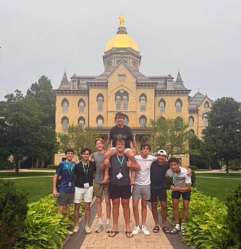 Eight smiling young men, one on shoulders, pose before Notre Dame's Main Building with its golden dome.
